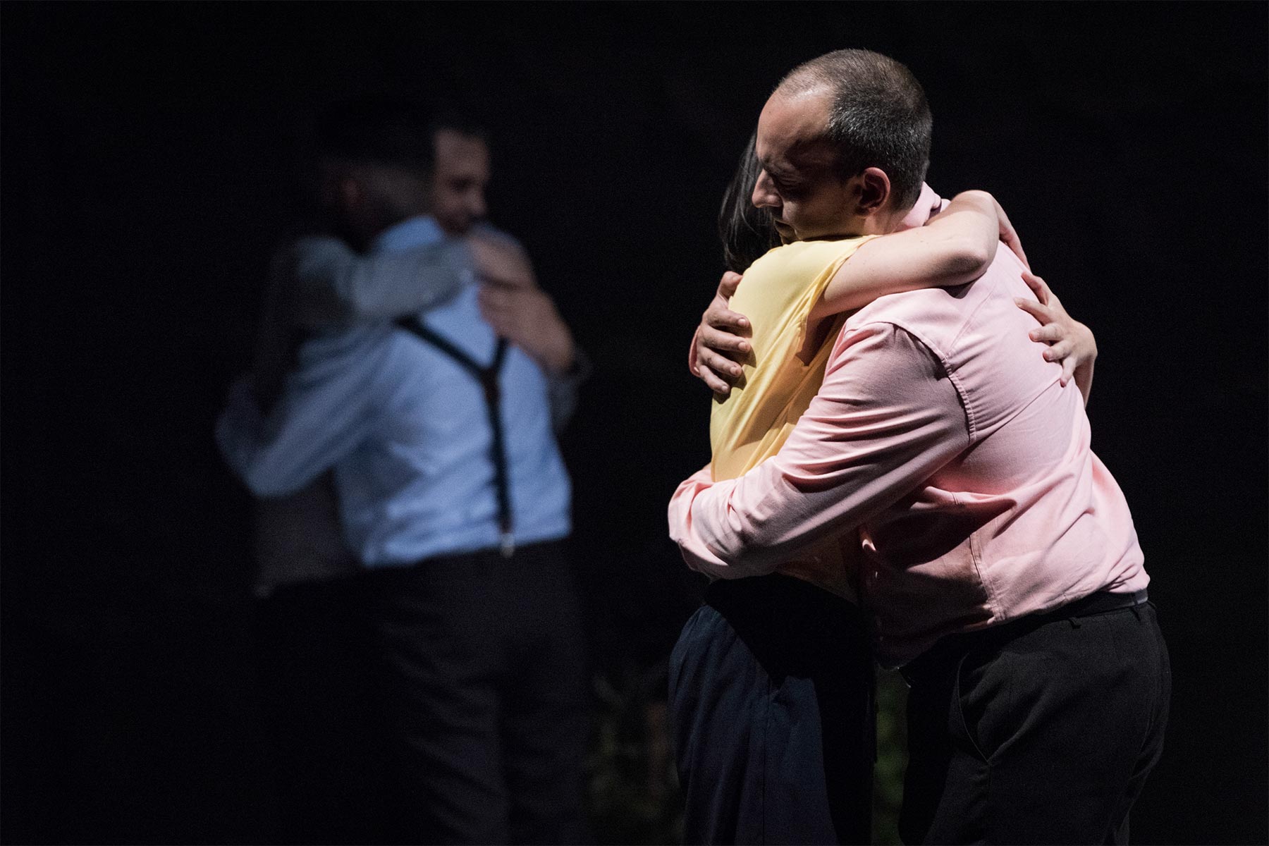 Persona abrazándose en una escena teatral, mostrando emociones intensas y cercanía, en un entorno oscuro que resalta la expresión y la conexión entre personajes.