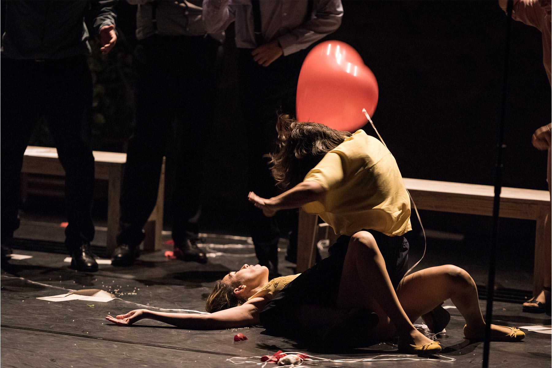 Una mujer en escena con un globo en forma de corazón, participando en una representación teatral expresiva, rodeada de personas en un escenario con elementos dramáticos y emocionales.