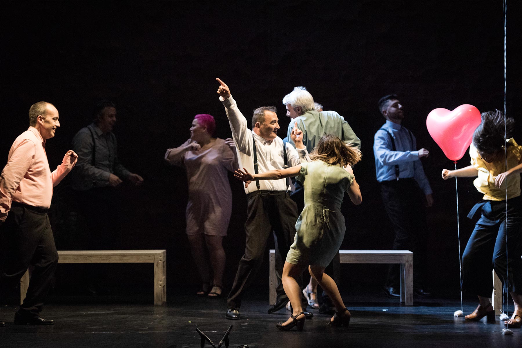 Varios actores en una obra de teatro representan una escena alegre con baile y globos en un escenario oscuro.