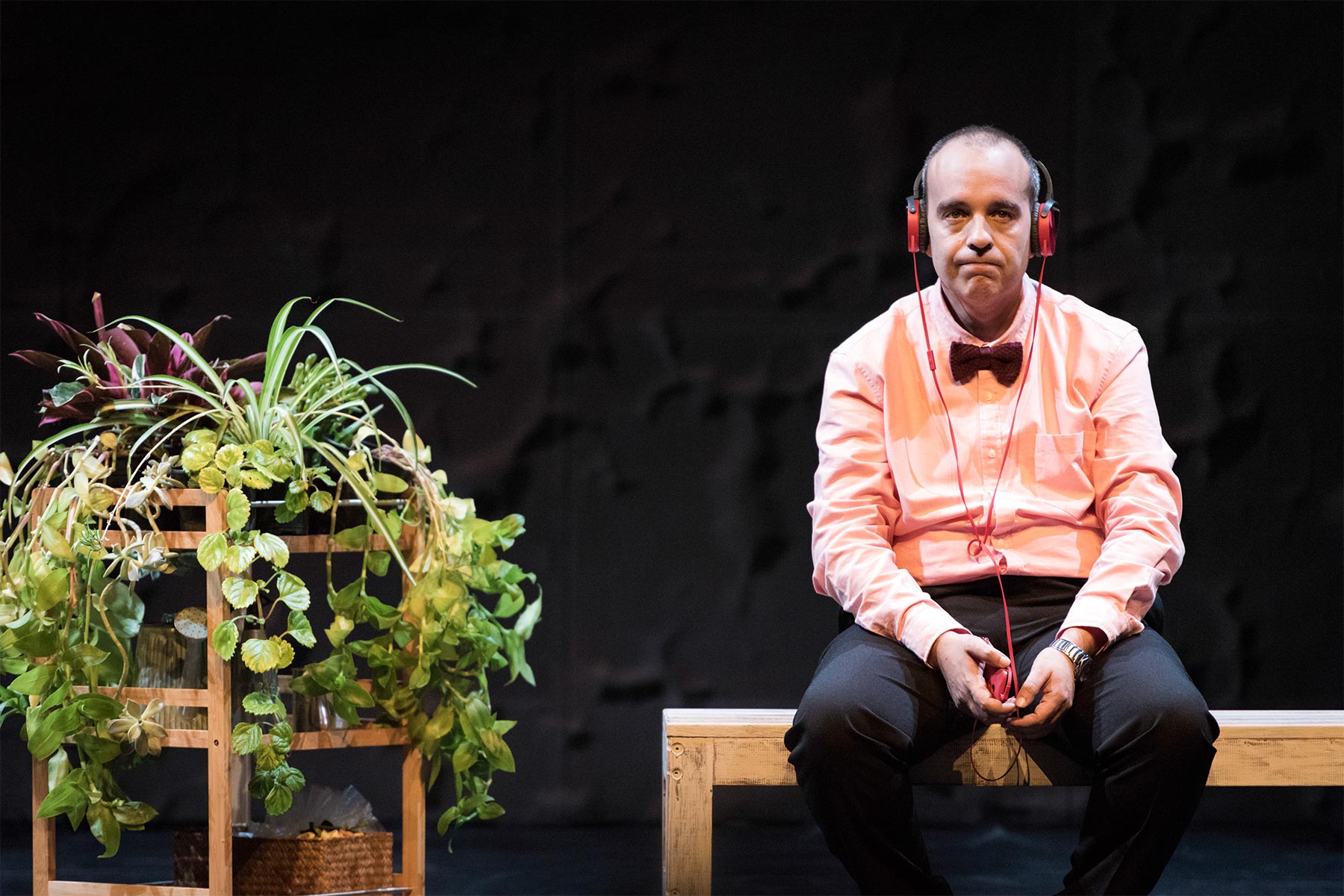 Boceto de un hombre con camisa rosa, pajarita y auriculares sentado en un banco de madera en un escenario teatral, expresando una emoción intensa, junto a un mueble con plantas decorativas, en un entorno de teatro profesional.