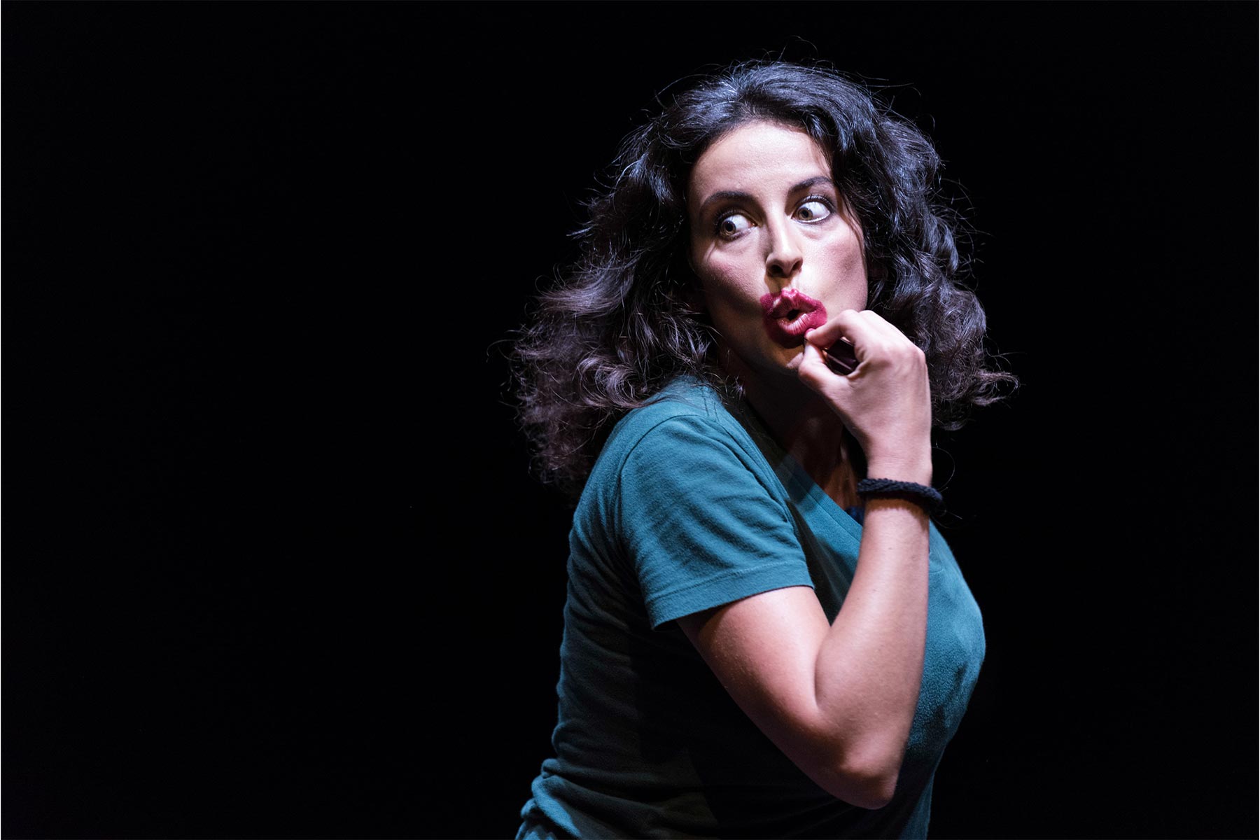 Andrea Jimenez en escena, mujer con cabello rizado y expresión pensativa, vestida con camiseta azul y fondo negro, mostrando su talento en artes escénicas y expresión artística.