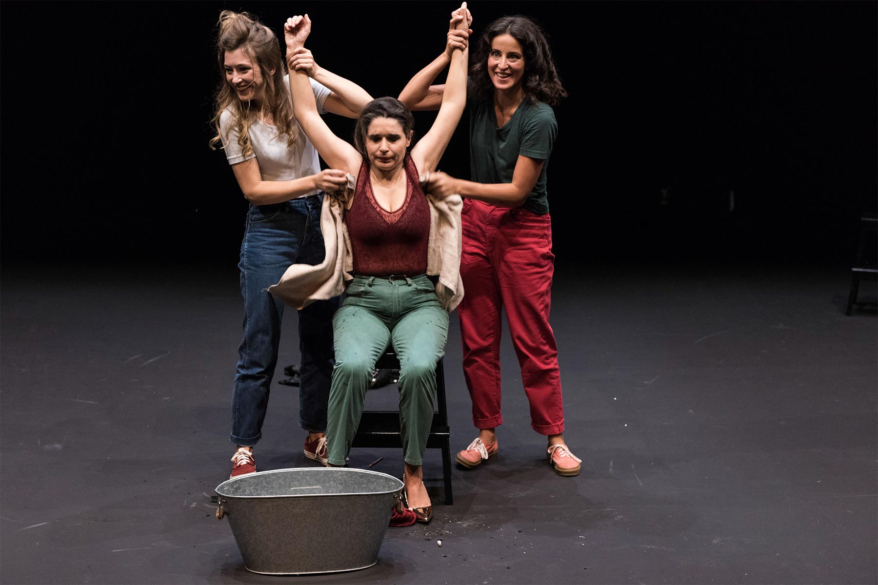 Bañista en teatro actuando con dos actrices en escena, expresión dramática, escenario oscuro y foco en la interpretación teatral, representando emociones intensas en una obra artística.