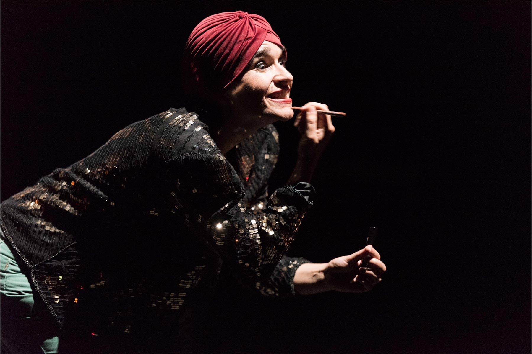 Maquilladora profesional maquillando en un escenario con vestido brillante y turbante rojo.