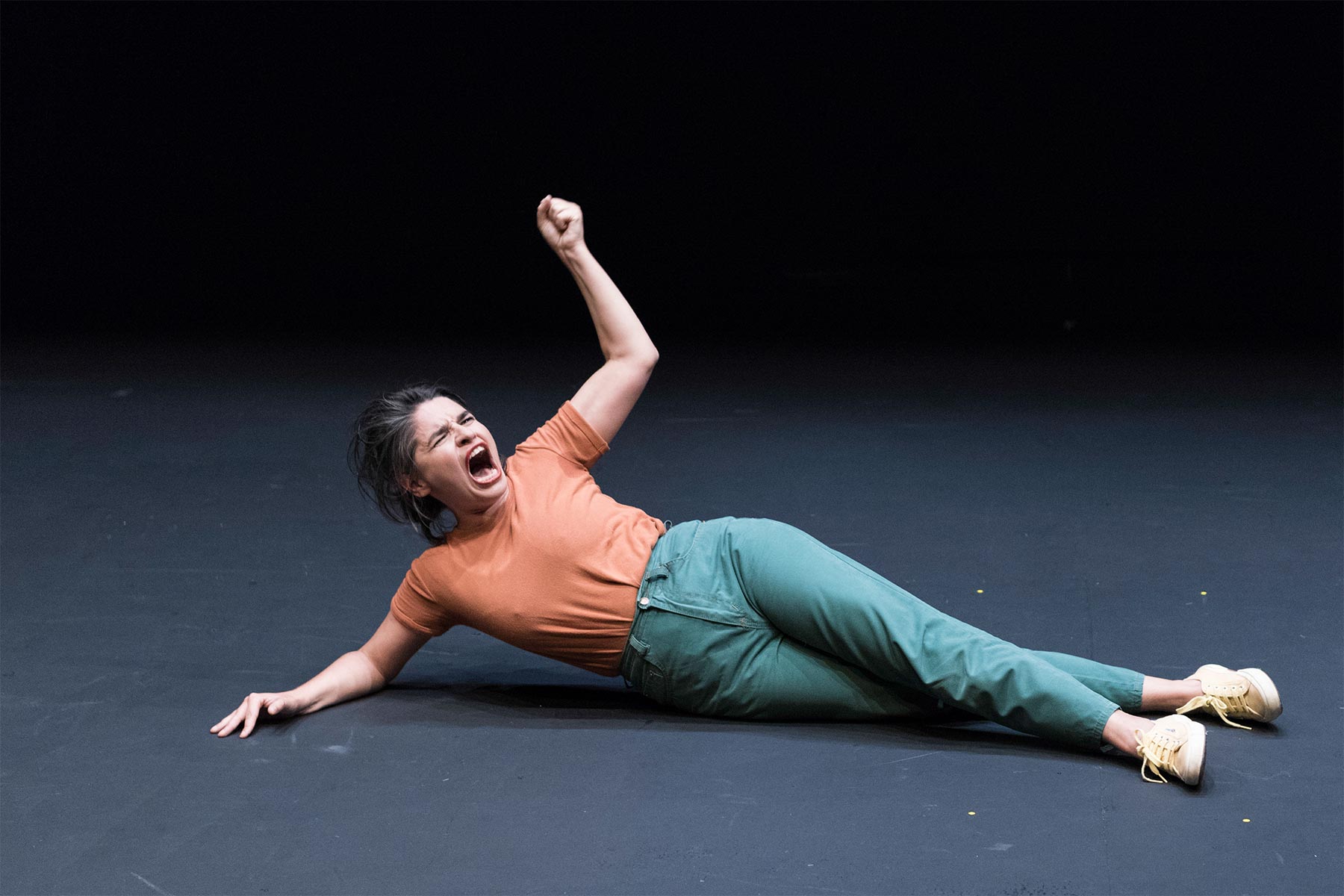 Grito de angustia de una mujer en el suelo, escena teatral con expresión de dolor, fondo negro y escenario oscuro, mostrando emociones intensas y actuación dramática.