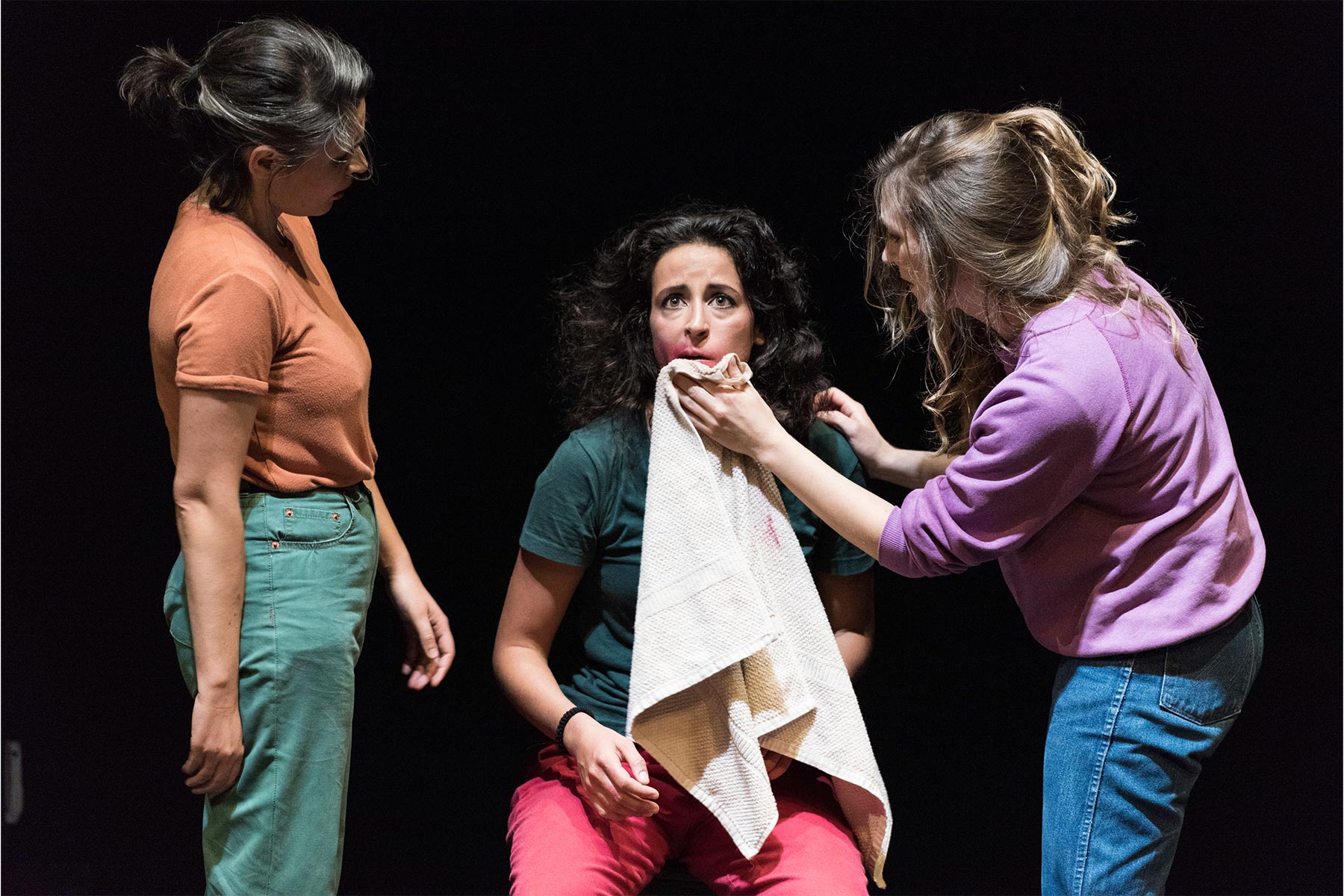 Novia en escena durante una obra de teatro, interpretando un papel dramático con expresiones intensas y gestos profundos, acompañada por dos actrices en un escenario oscuro, destacando por su interpretación emocional y presencia escénica.