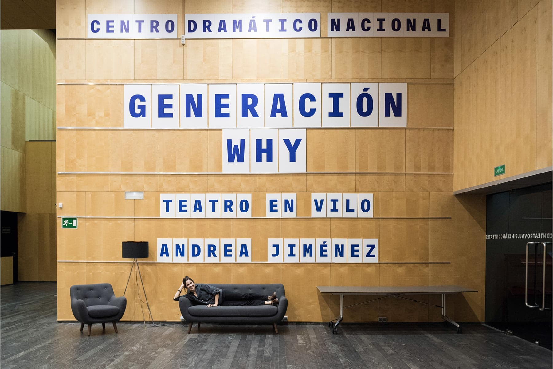 Innovadora mujer en el vestíbulo del Centro Dramático Nacional con cartel de "Generación Why" y "Teatro en Vilo" para promover eventos culturales y teatro en Madrid.