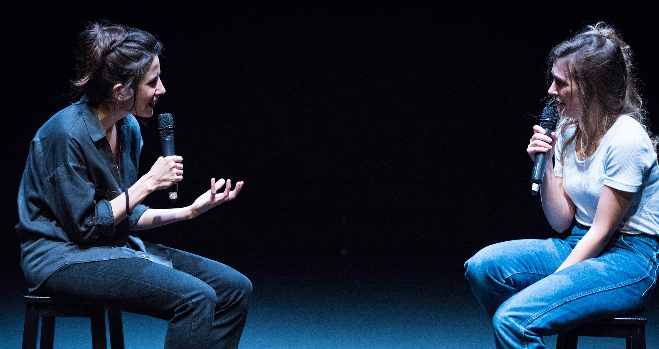 Diálogo entre dos jóvenes en una entrevista o conversación en un escenario, usando micrófonos, en un ambiente de teatro o conferencia.