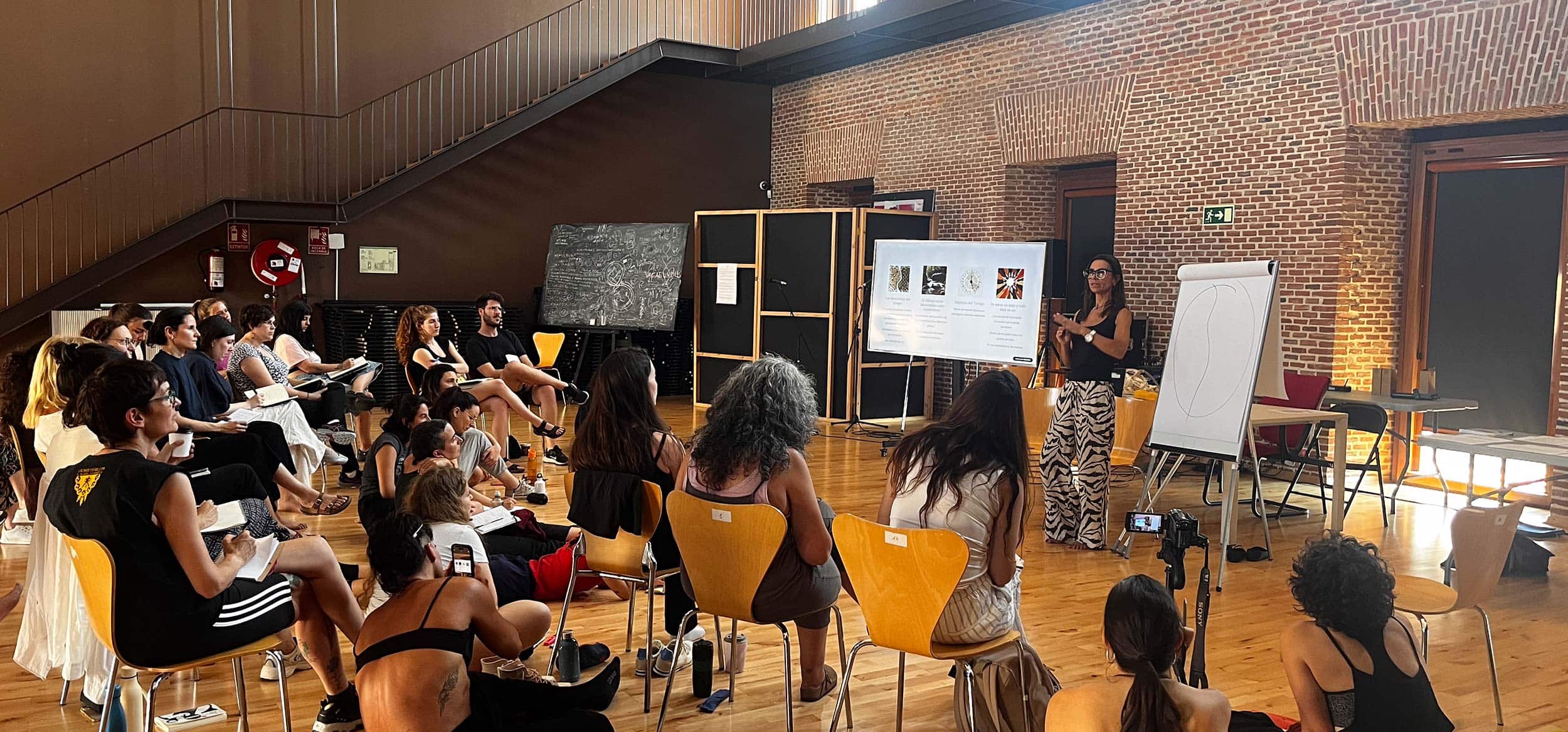 Imagen de una conferencia en un espacio amplio con asistentes atentos y una ponente explicando en la pizarra, en un entorno de estética moderna con paredes de ladrillo y suelo de madera.