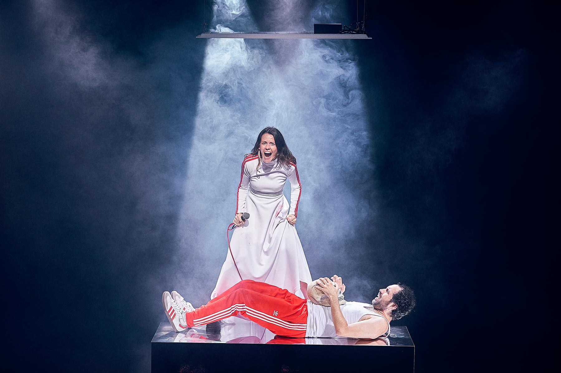 Disputa teatral en escena con actriz y actor en un escenario con efectos de humo y luz dramática.